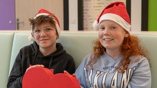 Ein Junge mit Brille und ein Mädchen mit Sommersprossen tragen Weihnachtsmützen und halten zusammen ein Herz.