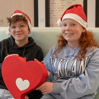 Ein Junge mit Brille und ein Mädchen mit Sommersprossen tragen Weihnachtsmützen und halten zusammen ein Herz.