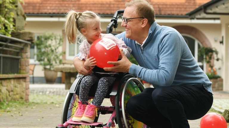 Mädchen im Rollstuhl hält einen roten Luftballon und lächelt den Vater an