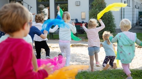 Kinder rennen zu einem Sandkasten. In den Händen halten sie bunte Jongliertücher.