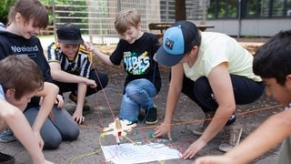 Mehrere Schülerinnen und Schüler einer Förderschule knien im Kreis um ein selbstgebautes Modell, das mit Schnüren verbunden ist.