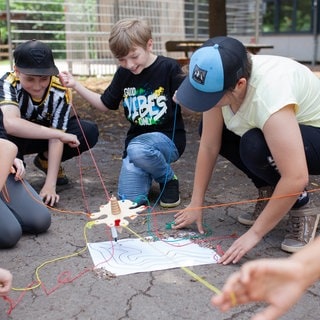 Mehrere Schülerinnen und Schüler einer Förderschule knien im Kreis um ein selbstgebautes Modell, das mit Schnüren verbunden ist.