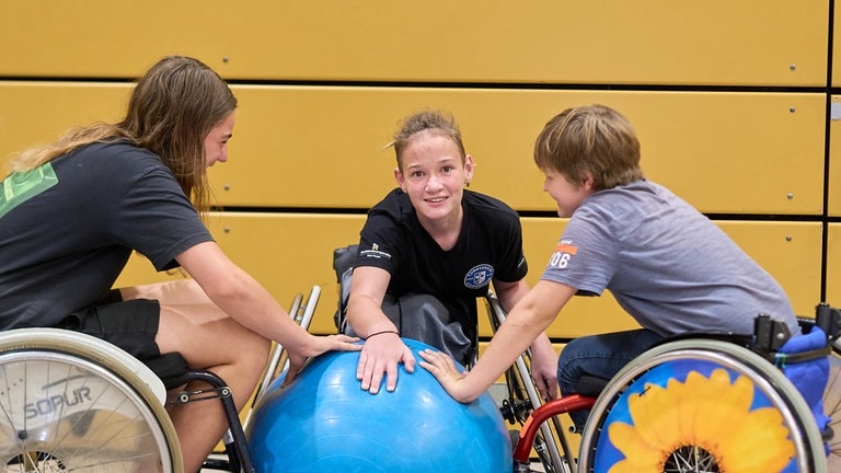 Drei Jugendliche in Rollstühlen halten ihre Hände auf einen blauen Gymnastikball.